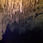 Strop z beżowych stalaktytów oświetlony latarką, odbijający się w czarnej tafli wody, w cenocie The Pit