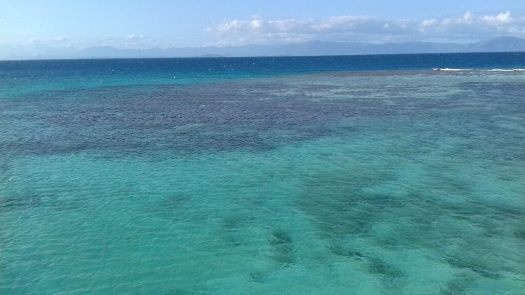 Laguna z góry, turkusowa woda z ciemnymi plamami koralowców, po prawej bariera, na której łamią się fale, a na horyzoncie góry Grande Terre