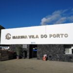 Budynek recepcji mariny w Vila do Porto: duży biały szyld z metalowymi literami na ścianie z czarnego bazaltu, pod błękitnym niebem, po lewej zielony klif