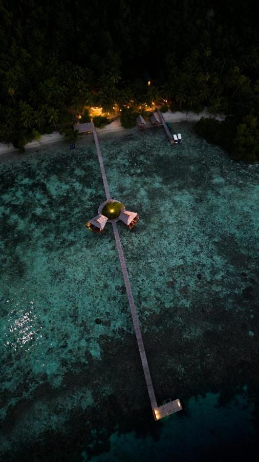 Nocny widok z lotu ptaka na długi drewniany pomost biegnący od plaży, z dwiema chatkami na palach w połowie drogi