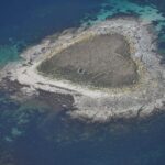 Widok z lotu ptaka na małą wysepkę z szarych płyt granitowych, porośniętą pośrodku suchą trawą, w której widać ruiny murków, z jasną plażą wokół, otoczoną turkusowymi płyciznami i ciemnoniebieską wodą.