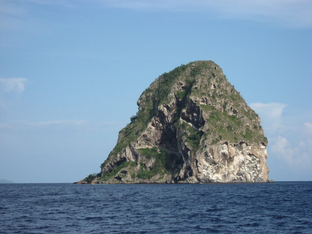 Rocher du Diamant z bliska od strony morza, masa szarej skały z jaskiniami, w białe smugi, z roślinnością na szczycie, nad ciemnoniebieskim morzem