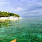 Przejrzysta turkusowa woda nad płytkim dnem, przed plażą z białego piasku obrzeżoną drzewami, przy której stoją w rzędzie białe łodzie bangka, a dalej inne łodzie pod zachmurzonym niebem.