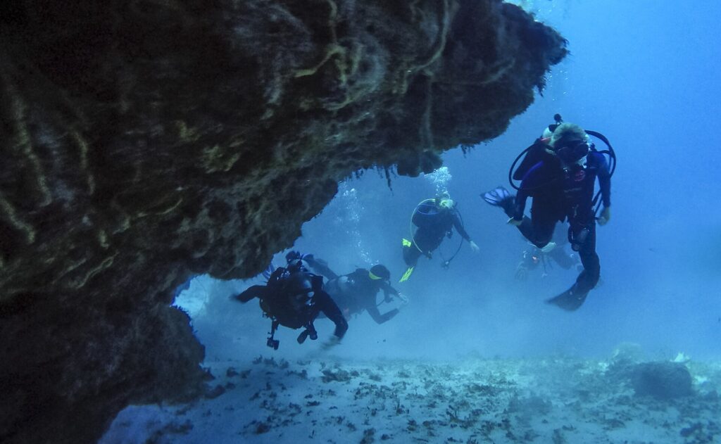 Czterech nurków wypływa z koralowego tunelu nad piaszczystym dnem na Cozumel, ze skalnym sklepieniem nad nimi