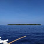Płaska, zalesiona wyspa Balicasag na horyzoncie, z białą latarnią morską i pasem jasnego piasku, widziana z łodzi bangka, której białe pływaki zajmują pierwszy plan, pod bezchmurnym błękitnym niebem.