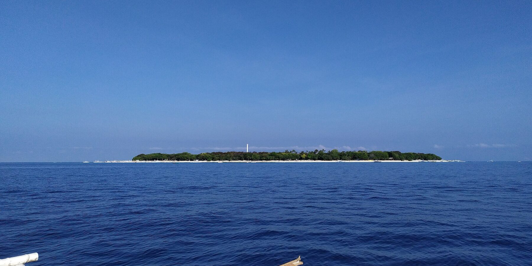 Płaska, zalesiona wyspa Balicasag na horyzoncie, z białą latarnią morską i pasem jasnego piasku, widziana z łodzi bangka, której białe pływaki zajmują pierwszy plan, pod bezchmurnym błękitnym niebem.