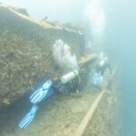 Dwaj nurkowie płyną wzdłuż pokładu Thistlegorm, przy korytarzu, w mlecznoniebieskiej wodzie