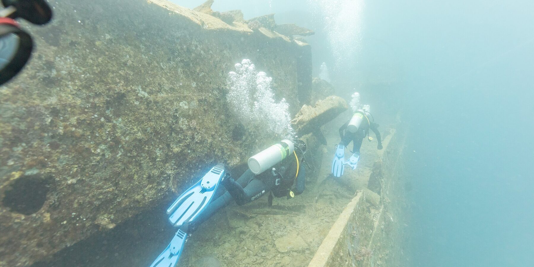 Dwaj nurkowie płyną wzdłuż pokładu Thistlegorm, przy korytarzu, w mlecznoniebieskiej wodzie