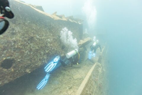 Dwaj nurkowie płyną wzdłuż pokładu Thistlegorm, przy korytarzu, w mlecznoniebieskiej wodzie