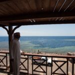 Z drewnianego tarasu mężczyzna w galabiji patrzy na nieckę Blue Hole otoczoną jasną rafą, w której pływają kąpiący się ludzie